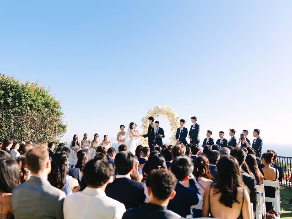 Outdoor wedding ceremony under a clear blue sky. A couple stands by a floral arch, surrounded by bridesmaids and groomsmen, with guests seated.