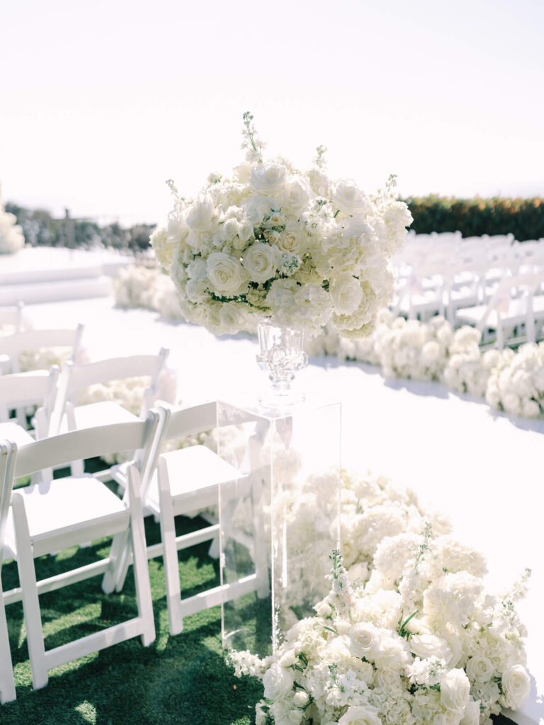 Outdoor wedding venue with rows of white chairs, floral arrangements, and an arch of white flowers. Overlooks a scenic, sunny ocean view. 