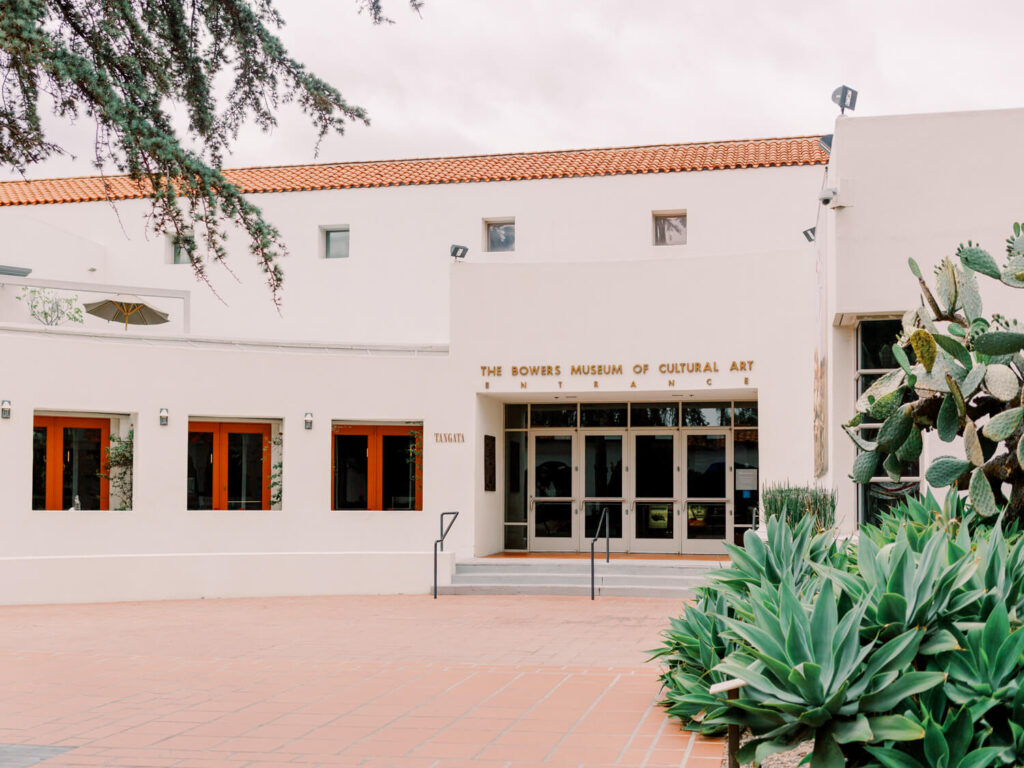 White building of Bowers Museum with a terracotta roof. Entrance flanked by plants and cacti.