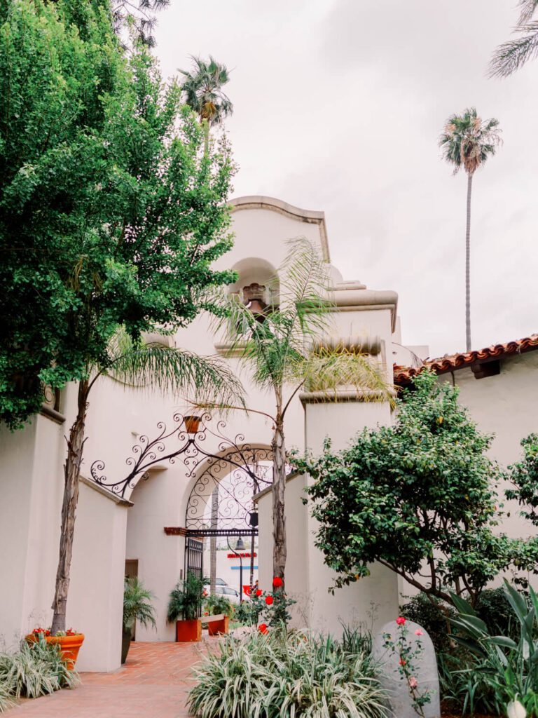 Spanish-style architecture with a stucco archway and ornate wrought iron details, surrounded by palm trees and lush greenery under an overcast sky.