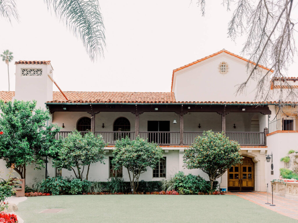 Spanish-style building with a red-tiled roof and second-floor balcony, surrounded by lush green trees and grass.