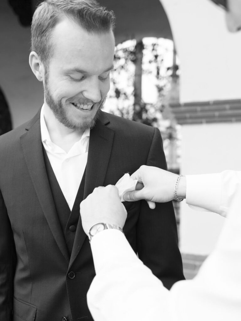Black and white image of a smiling man in a suit having a boutonniere pinned on his lapel by another person.