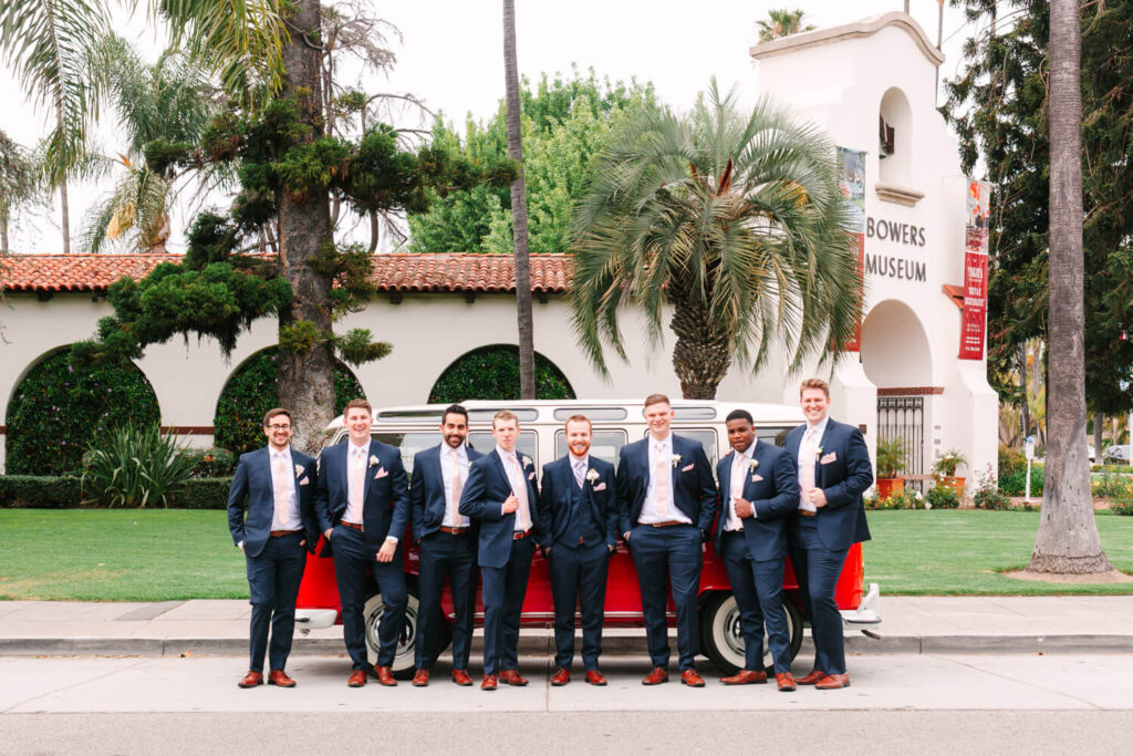 A group of eight men in matching navy suits and brown shoes stand casually in front of a red vintage van. They are in front of the Bowers Museum, surrounded by palm trees and greenery.