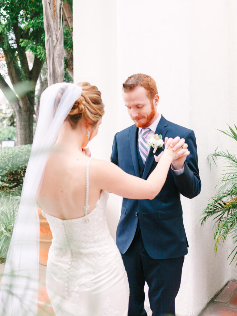A bride and groom share a tender moment outdoors, holding hands. The bride wears a lace dress and veil, the groom a navy suit.