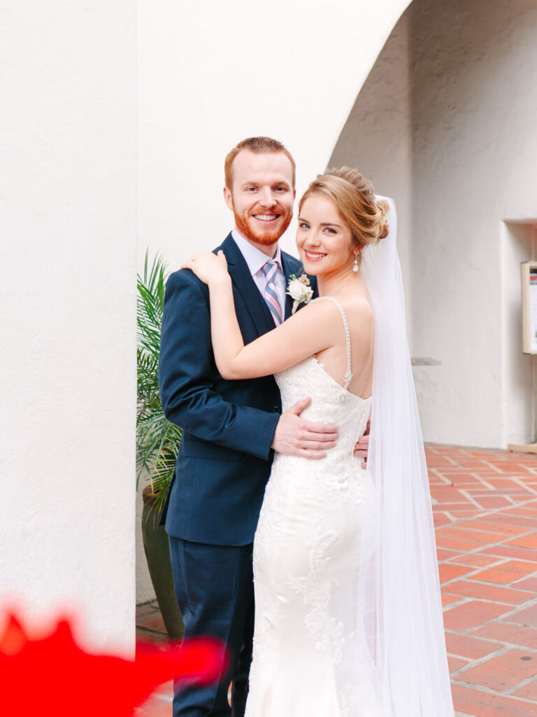 A smiling bride in a white dress and veil embraces a groom in a navy suit against a soft white wall.