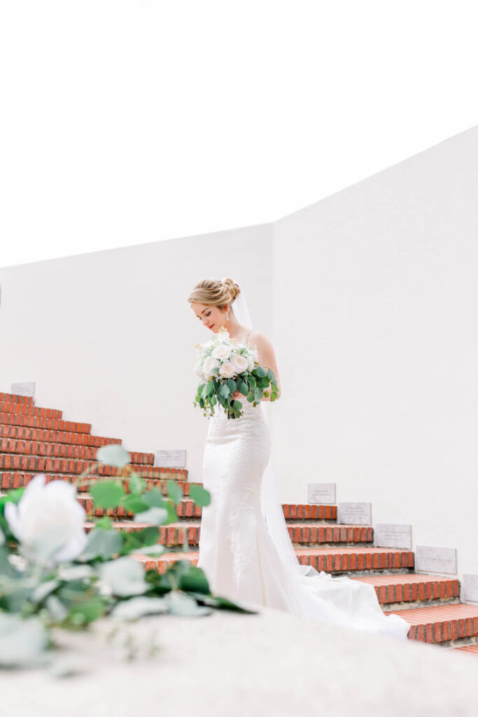 Bride in an elegant white dress descends a brick staircase holding a bouquet.