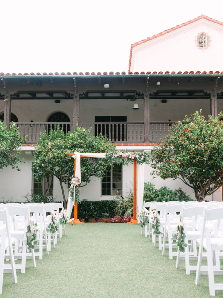 An outdoor wedding aisle with white chairs adorned with greenery leads to a floral arch. Two trees flank the arch, and a building with arched windows is in the background.