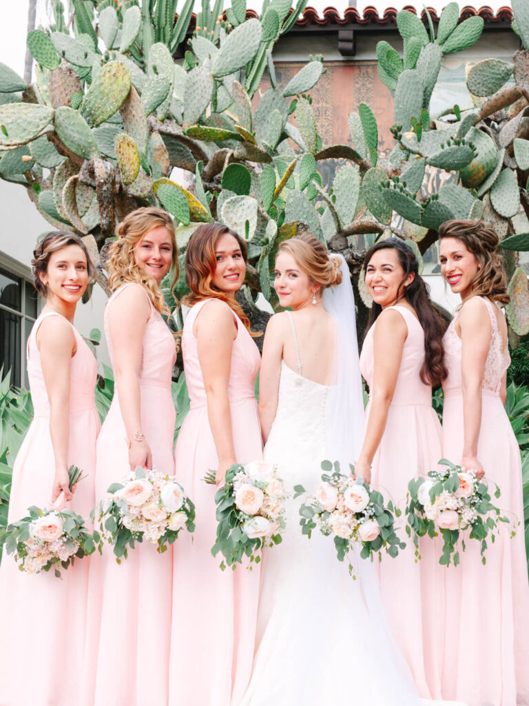 Bride in a white gown with five bridesmaids in blush pink dresses, holding bouquets. They stand joyfully in front of a large cactus plant.
