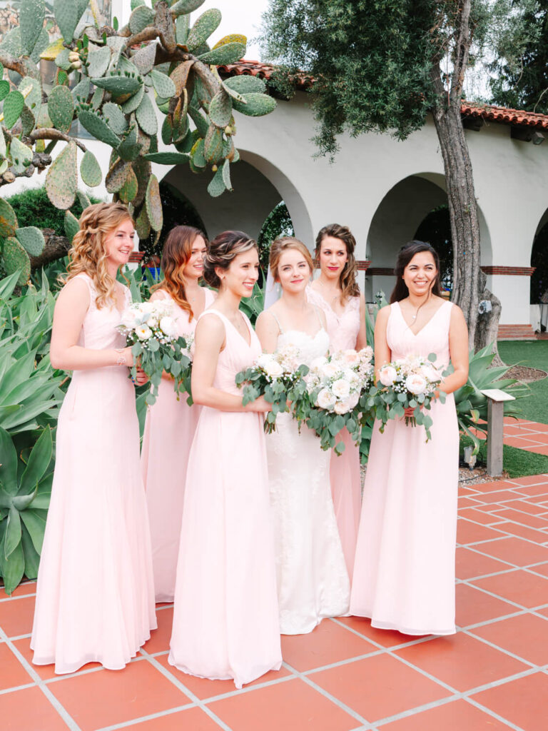 Bride stands with five bridesmaids, all in blush pink dresses, holding white floral bouquets. They're outdoors, surrounded by lush greenery and archways.