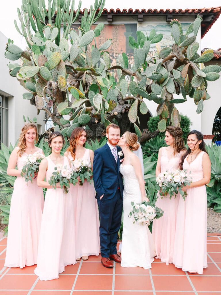 Bride and groom stand lovingly with six bridesmaids in pale pink dresses holding bouquets. A large cactus adds a rustic charm in the background.