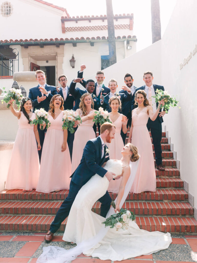 A joyful wedding party on steps. Bride in a white gown and groom in a navy suit share a romantic dip. Bridesmaids in pink; groomsmen in navy cheer with flowers.