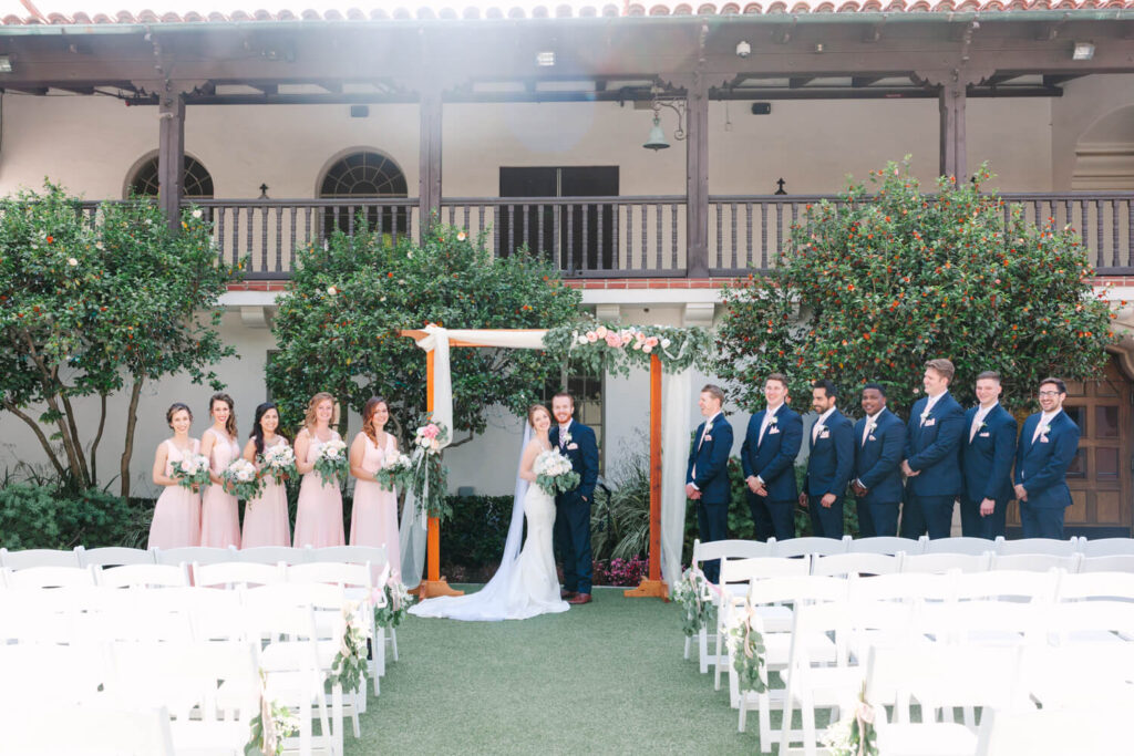 Bride and groom standing under a floral arch, surrounded by bridesmaids in pink dresses and groomsmen in blue suits, in a sunny outdoor courtyard.