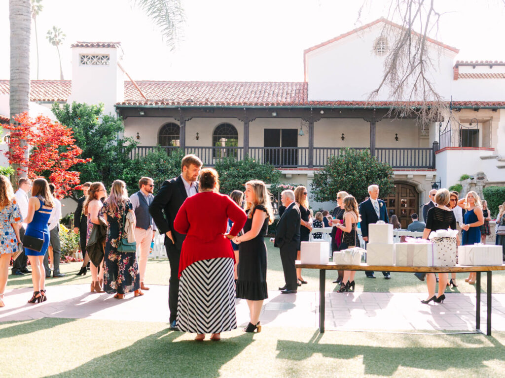 Outdoor garden party with guests in formal attire mingling under bright sunlight. A historic building with arched windows is in the background.