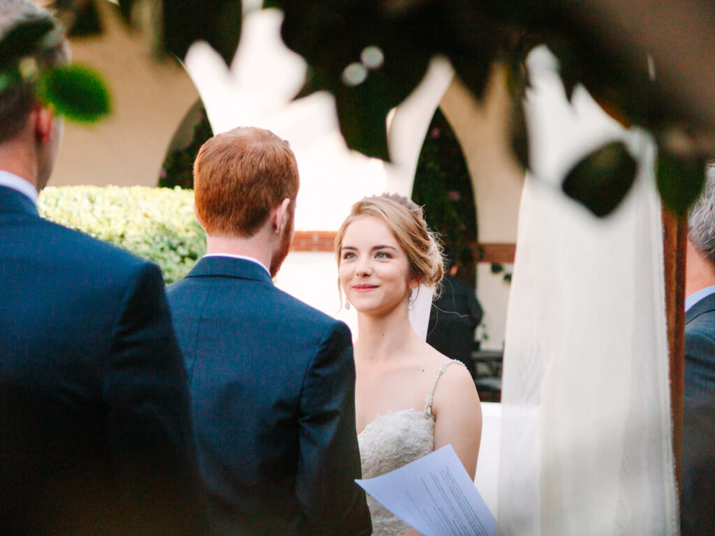 A bride in a white lace dress gazes affectionately at her groom during an outdoor wedding. The groom wears a dark suit, surrounded by soft foliage.