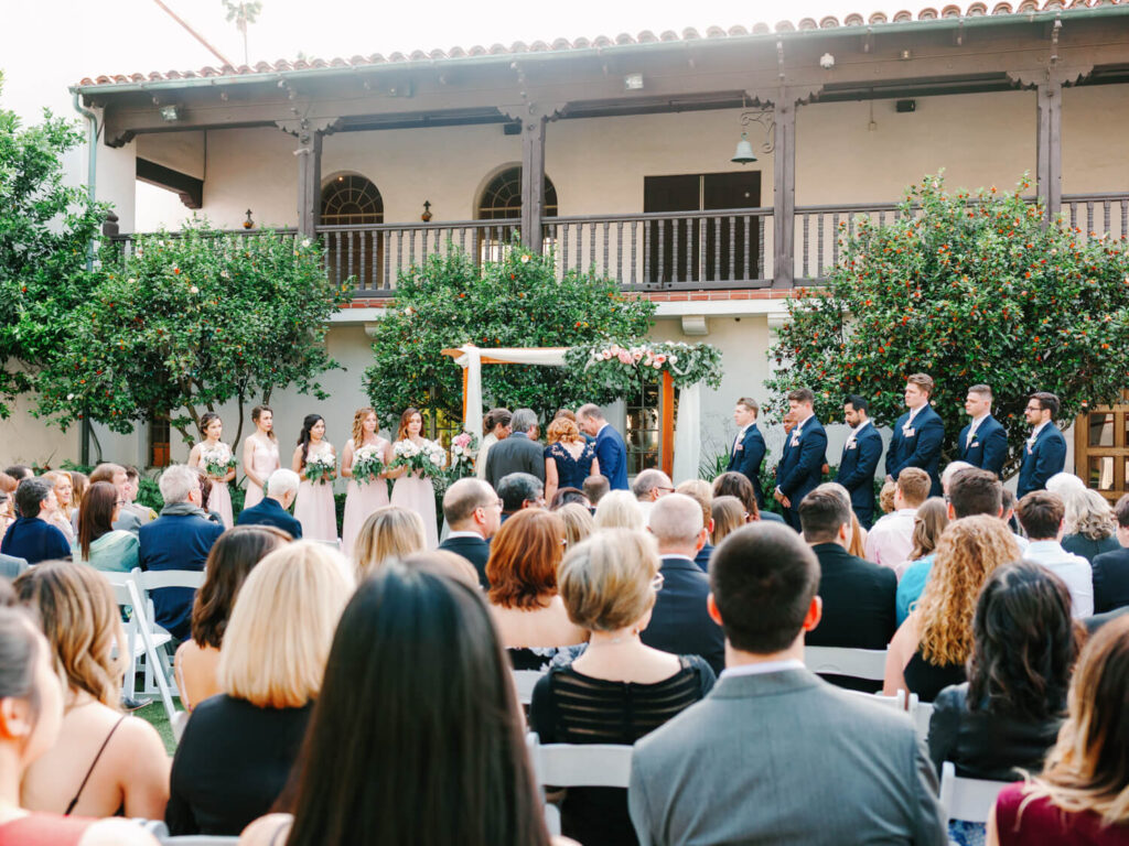 Outdoor wedding ceremony in a sunny courtyard. Bridesmaids in pastel dresses and groomsmen in suits stand near a floral arch, guests seated attentively.
