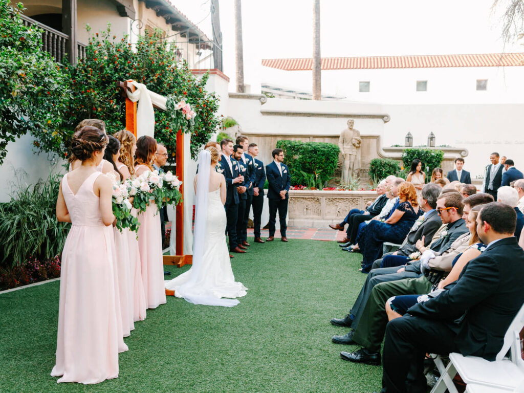 Bride and groom stand under a floral arch, facing each other. Bridesmaids in pink dresses hold bouquets to the left, with suited groomsmen on the right. Guests sit on white chairs on the green lawn.