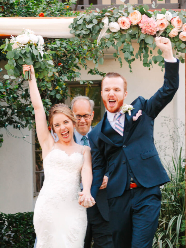 Bride and groom in wedding attire joyfully celebrate under floral arch, raising arms triumphantly. A smiling officiant stands behind them.