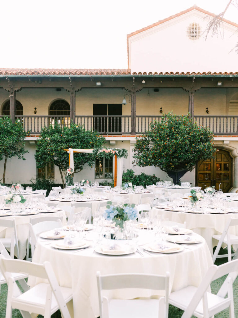 Elegant outdoor wedding setup in a courtyard with white round tables, white chairs, floral centerpieces, and a decorative arch, surrounded by greenery.