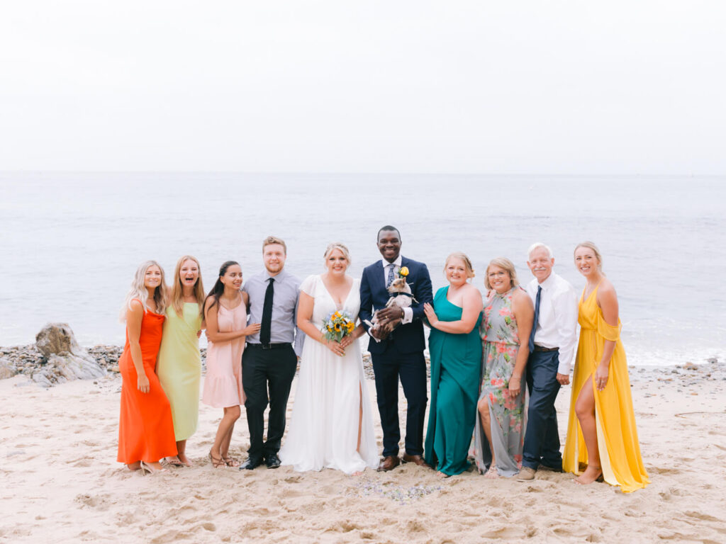A joyful wedding party poses on a sandy beach with the ocean in the background. The group includes the bride in a white dress, the groom in a dark suit, and nine smiling guests in colorful attire.