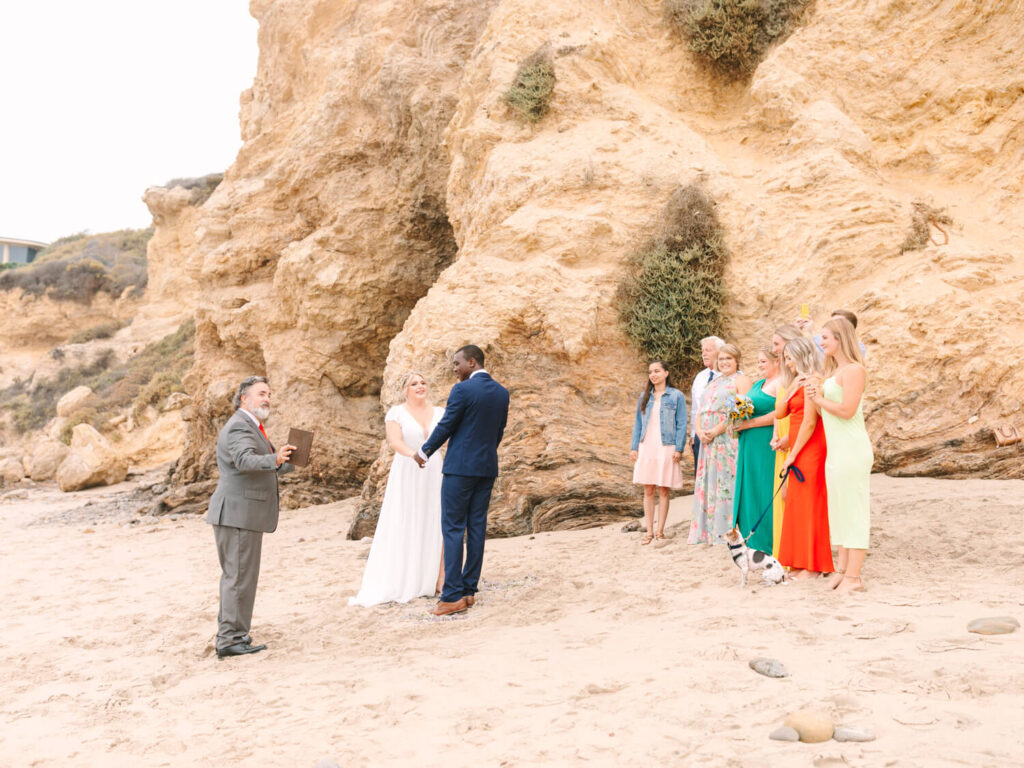 A beach wedding with a couple holding hands, officiant nearby. Guests, wearing colorful outfits, stand by a rocky cliff under a clear sky, creating a joyful, intimate atmosphere.