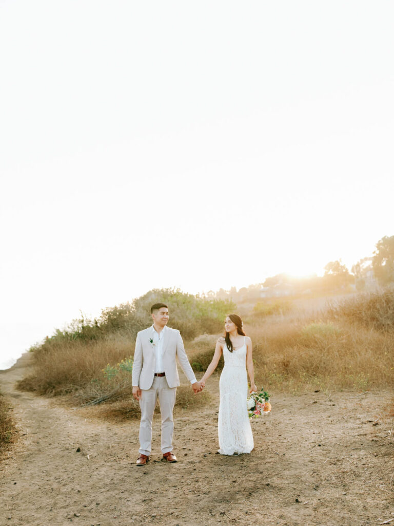 A couple holds hands in a sunlit field, the woman in a white lace dress holding a bouquet, while the man wears a light suit.