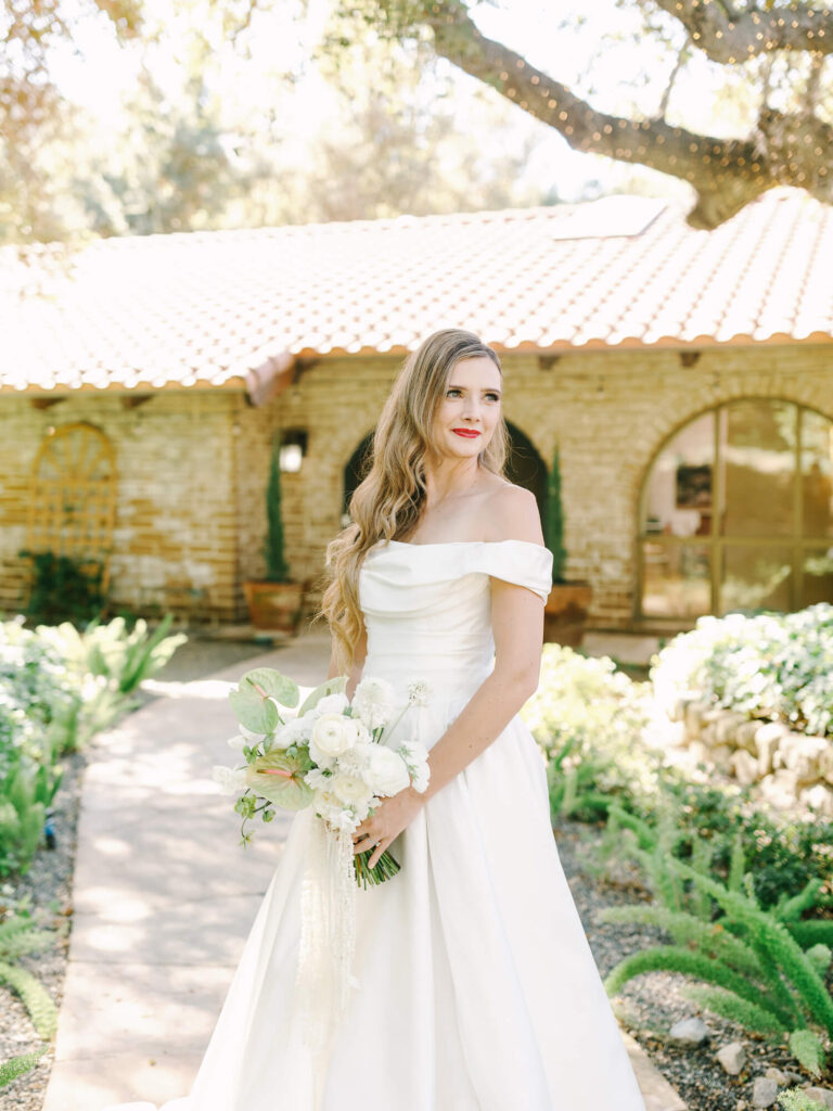 Bride in an off-shoulder white gown holding a bouquet of white flowers stands outside a rustic house with arched windows, surrounded by greenery.