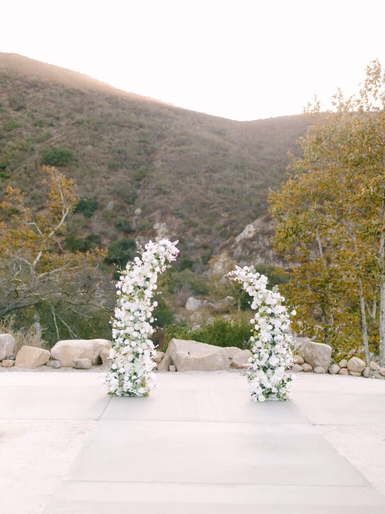 Two white floral arches on a stone patio, framed by rocky hills and autumn trees.