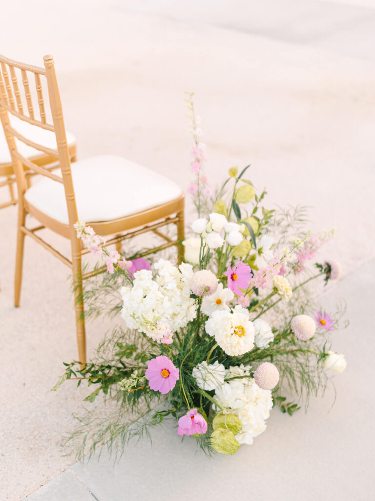 A wooden chair with a white cushion is beside a vibrant floral arrangement, featuring pink, white, and yellow blooms.