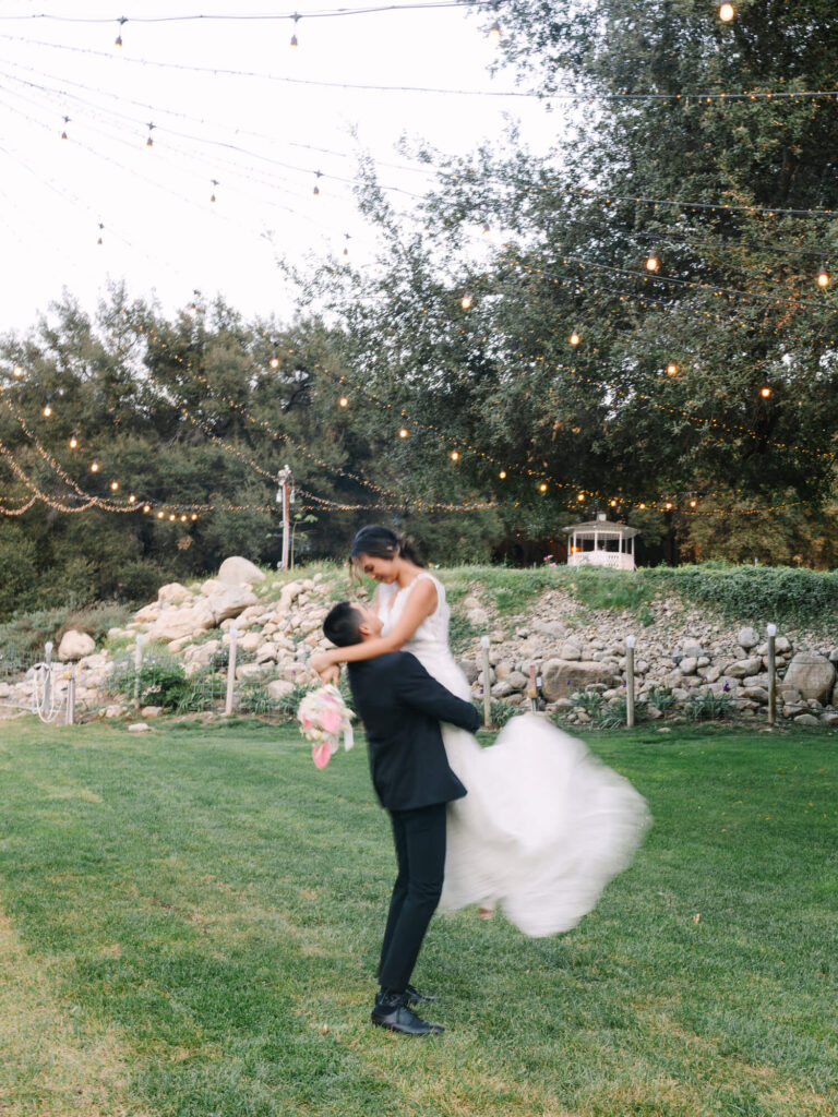 A groom lifts the bride playfully on a lush green lawn under string lights and trees, conveying joy and romance. She holds a pink bouquet.