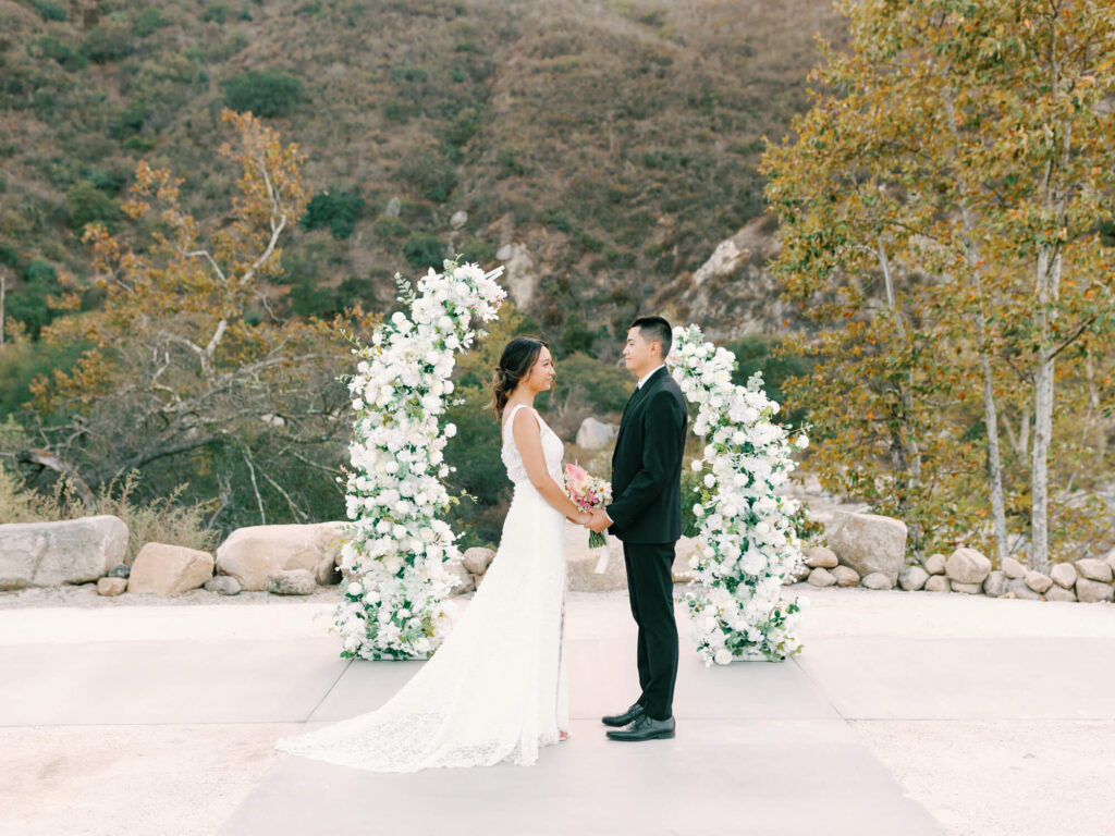 Bride and groom stand facing each other outdoors, surrounded by lush greenery and two elegant floral arches.