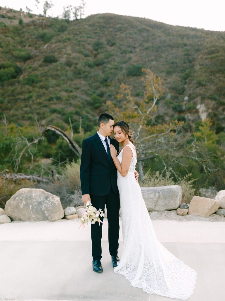 A bride and groom embrace tenderly outdoors, set against a backdrop of lush green hills. The bride wears a white gown, and the groom holds a bouquet.