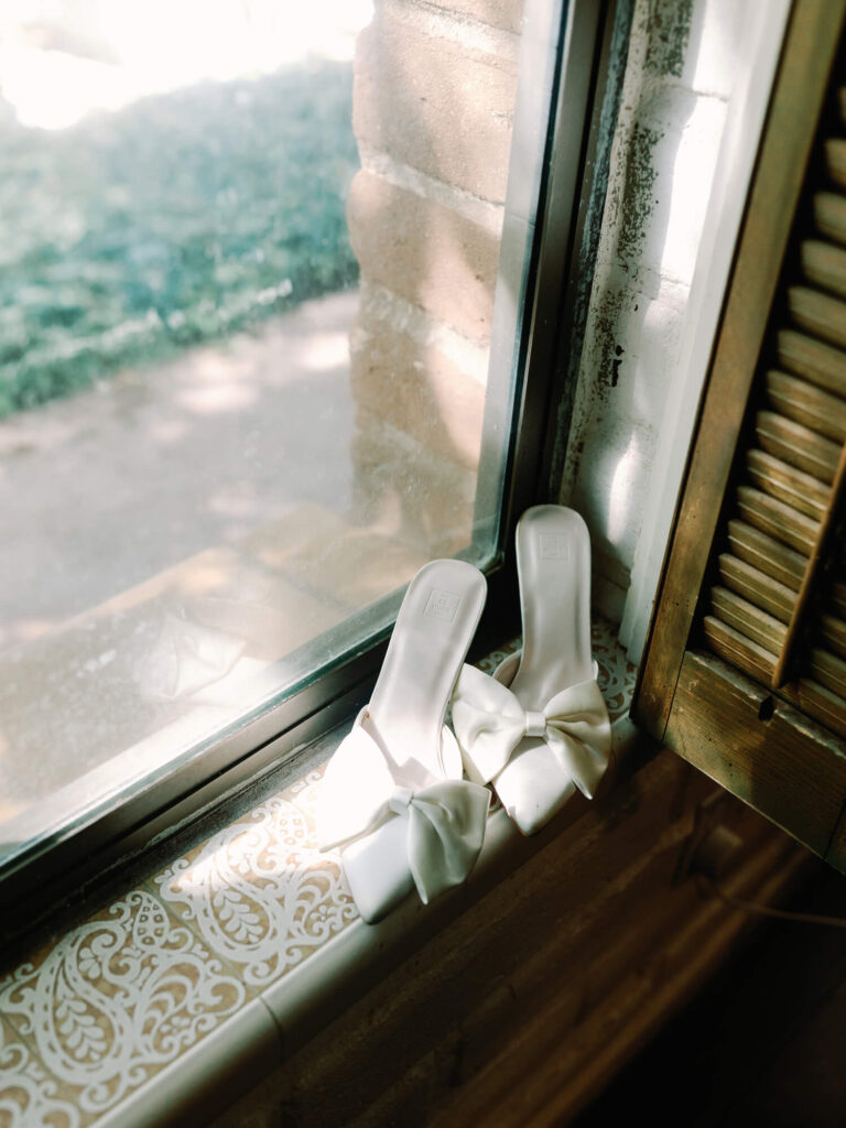 White satin mule heels with large bows rest on a tiled windowsill, bathed in soft, natural light.