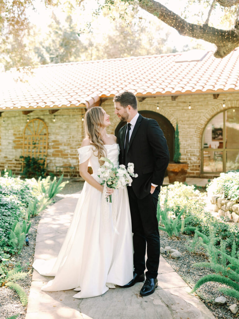 Bride in a white off-shoulder gown and groom in a black suit smile at each other outdoors. They stand on a garden path, with a rustic building behind them.