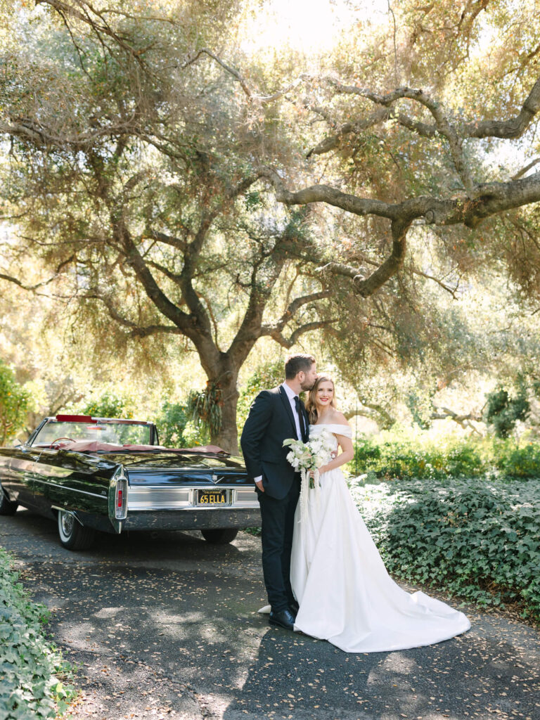 Bride and groom stand lovingly under a large tree beside a vintage black car.
