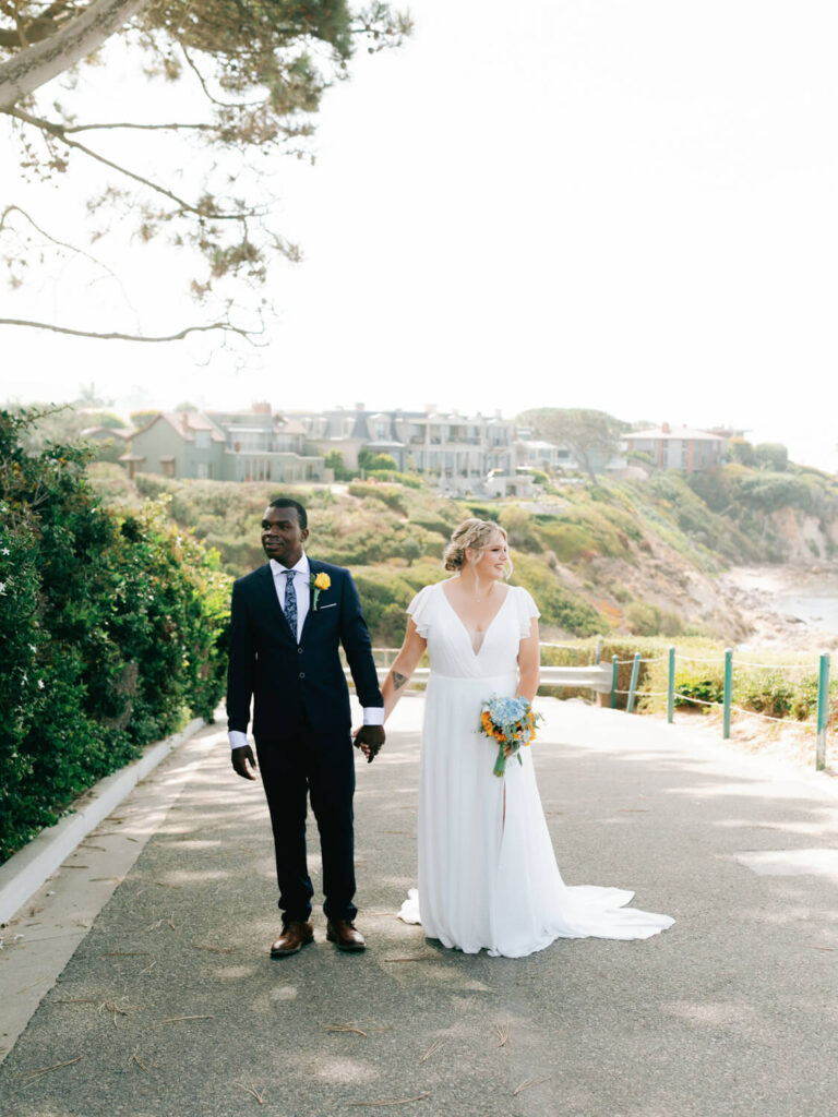 A bride and groom are holding hands on a sunlit path. She wears a white dress and holds a vibrant bouquet, while he wears a dark suit with a yellow boutonniere. Coastal cliffs and houses form the scenic background.