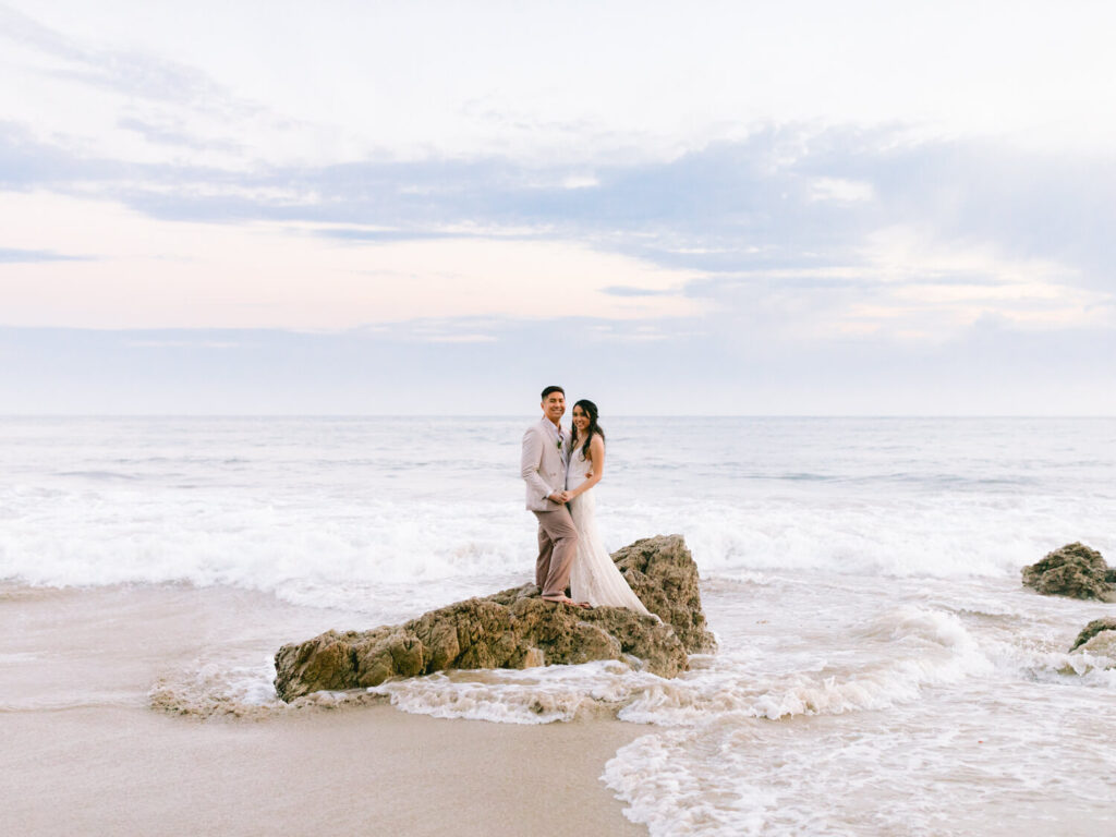 A couple stands on rocks at a beach, surrounded by gentle waves under a pastel sky.