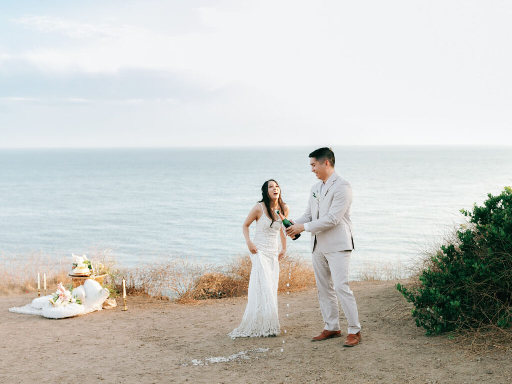 A joyful couple in wedding attire stands by the ocean. The man opens a champagne bottle as the woman laughs. A picnic setup is visible nearby.