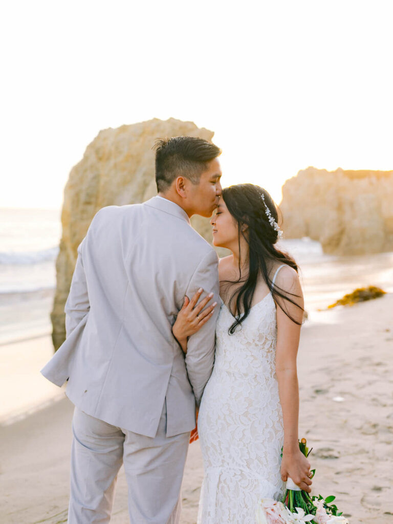 A couple embraces lovingly on a beach at sunset. The groom kisses the bride's forehead, both wearing elegant wedding attire.