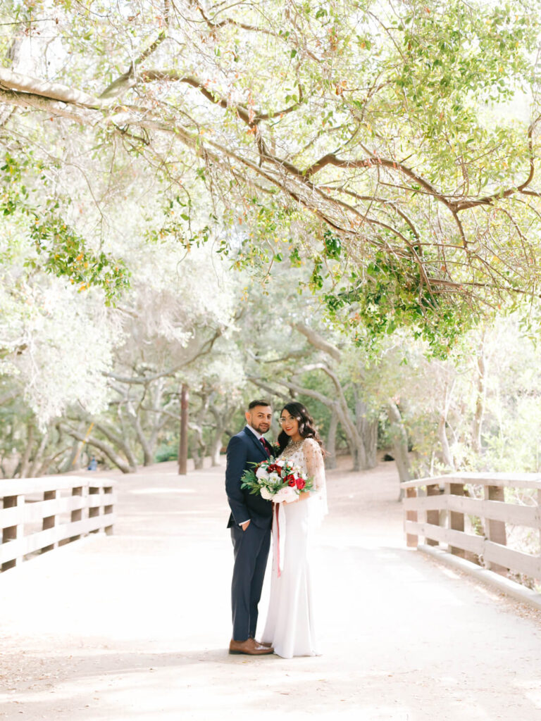 A couple stands on a sunlit wooden bridge beneath lush, arching branches. The bride in a white gown holds a bouquet, exuding joyful warmth and elegance.