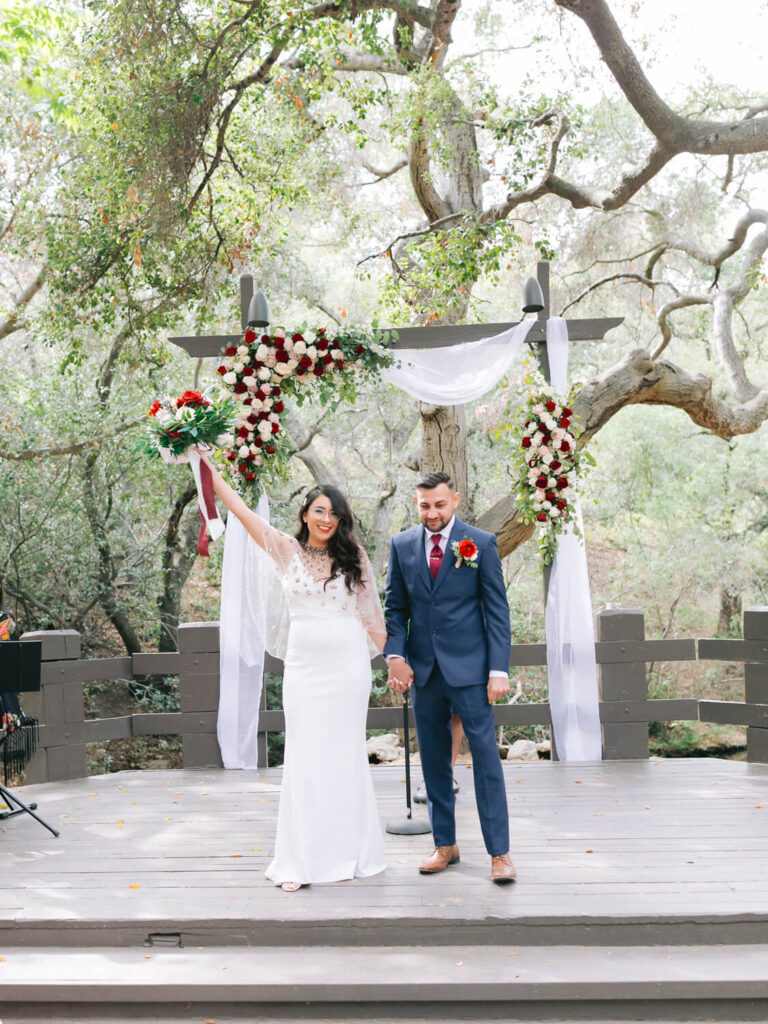 Bride in a white gown and groom in a navy suit stand under a floral arbor, with greenery background. She holds a bouquet high, smiling joyfully.