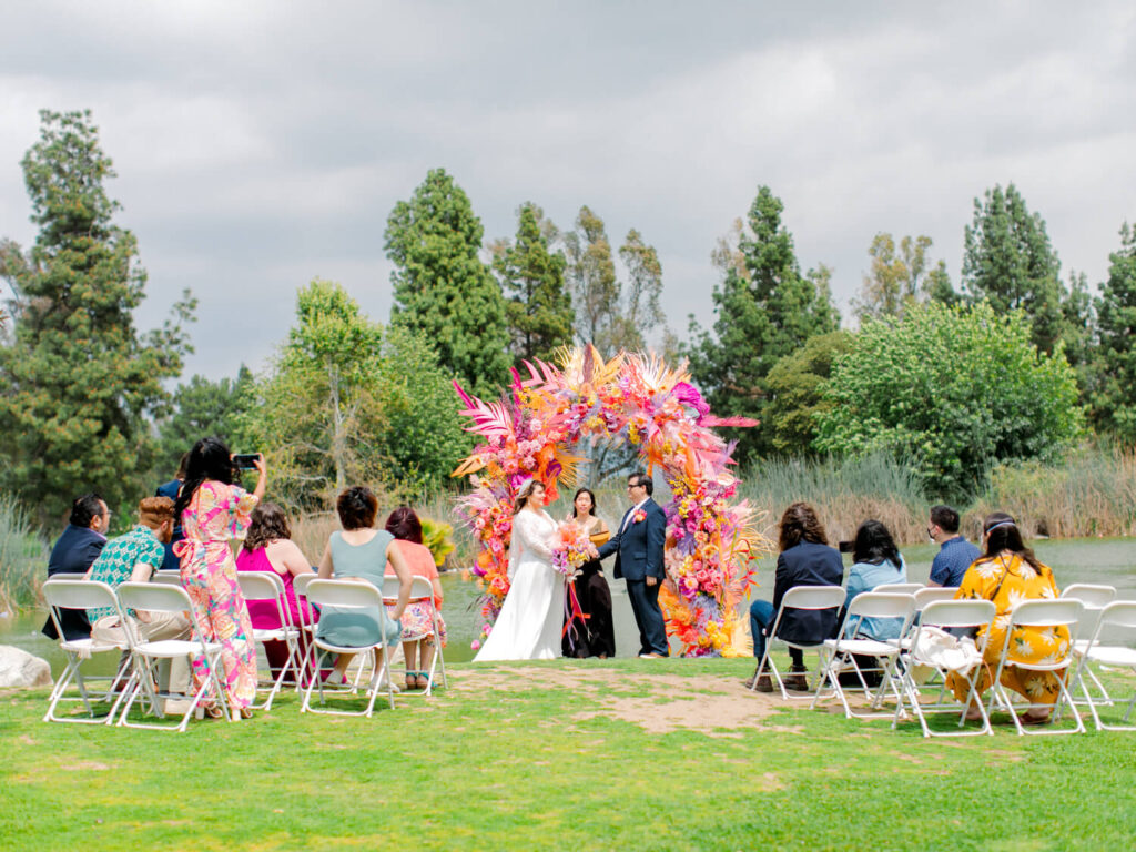 A couple stands at the altar in front of a vibrant floral arch during an outdoor wedding. Guests sit on white chairs on a grassy area, with a serene lake and lush trees as the backdrop.