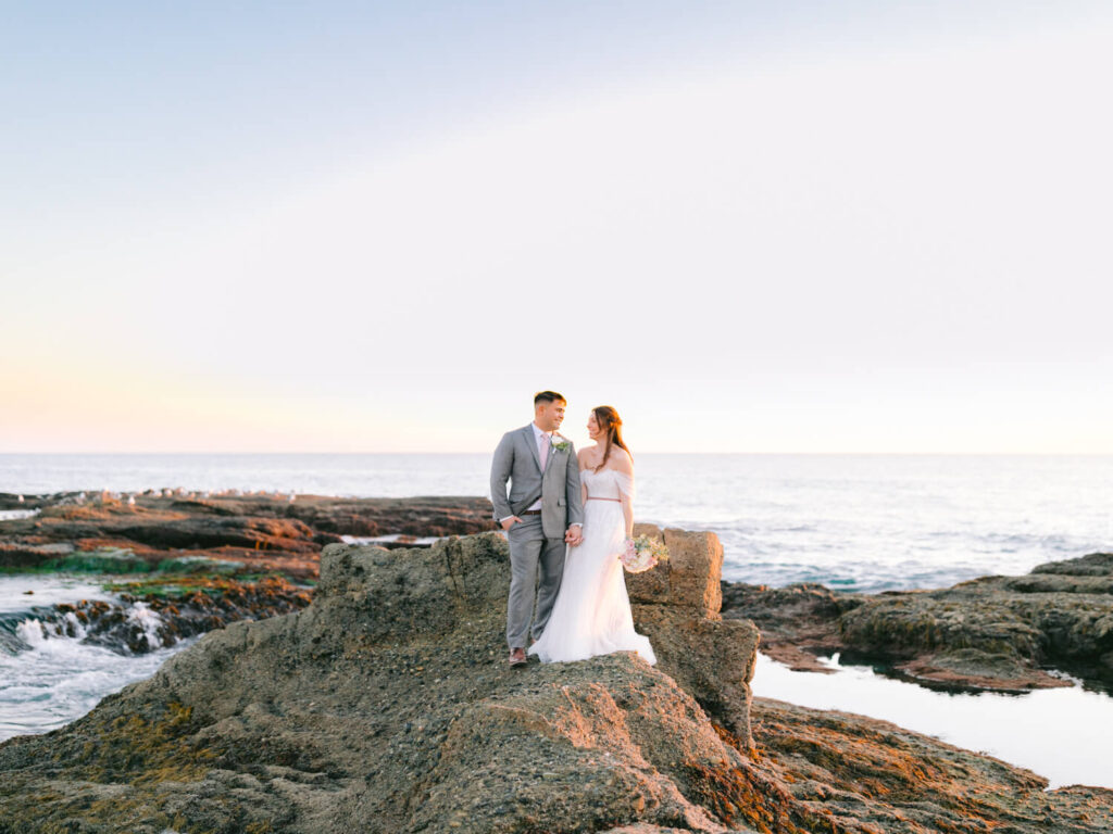 A bride in a white gown and groom in a gray suit stand on rocky shore at sunset, holding hands. Ocean waves crash gently in the background.
