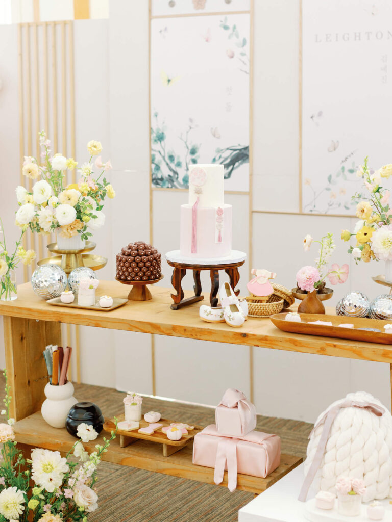 Elegant dessert table with a white cake adorned with pink ribbons, surrounded by pastel flowers, sweets, and decor in a soft and celebratory setting.