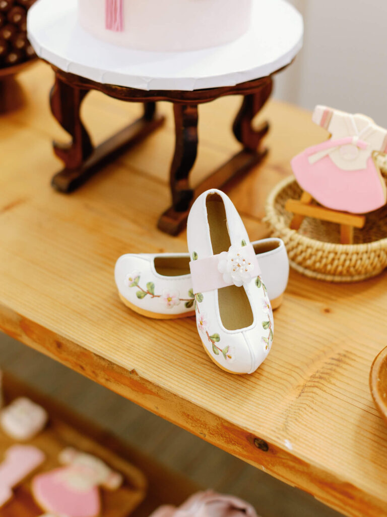 White, floral-decorated baby shoes rest on a wooden table near a pink cake. Beside them, a small pink dress cookie adds a playful, sweet touch.