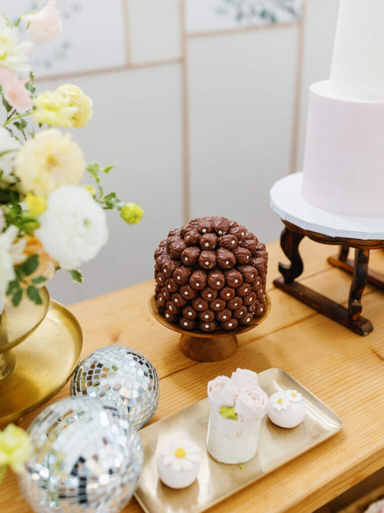 Elegant dessert table with a chocolate cake adorned with pearls, a floral arrangement, metallic orbs, and a cake with white and pink tiers.