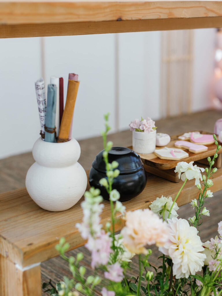A cozy wooden shelf displays a white vase holding rolled papers, a small black pot, and a tray with pink flowers and pastel cookies. Soft flowers add a fresh, serene touch.