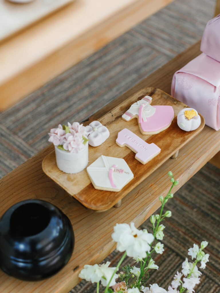 A wooden tray holds pink and white treats shaped like a dress, cross, and flower. Nearby, a pink bag and white flowers evoke a delicate, celebratory tone.