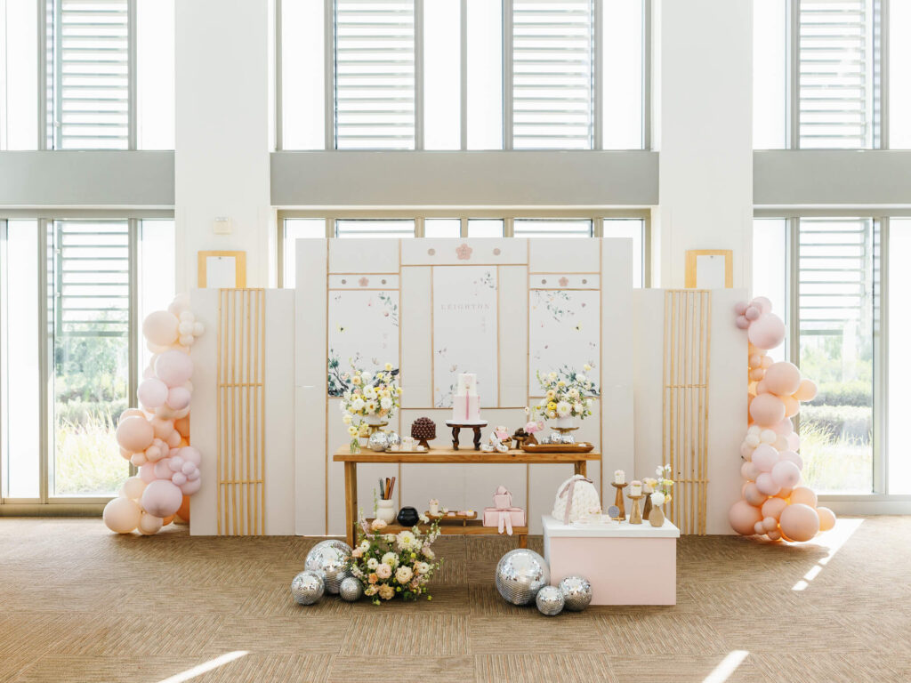 Elegant dohl party setup with pastel balloons, floral arrangements, and decorative sweets on a wooden table. Soft, natural light streams through large windows.