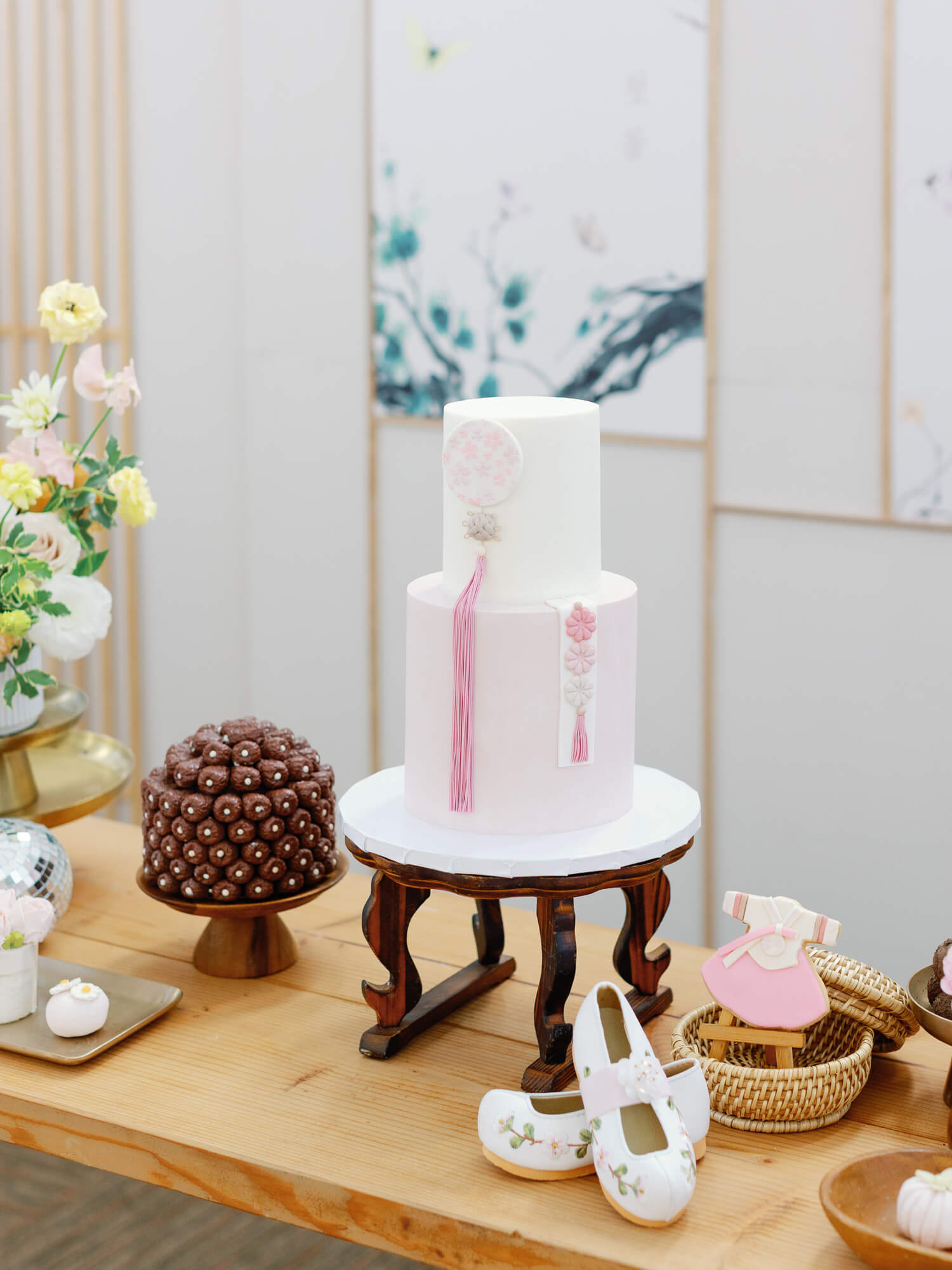 Two-tier cake with pink floral designs and tassels on a wooden table. Nearby are floral baby shoes and chocolate balls.