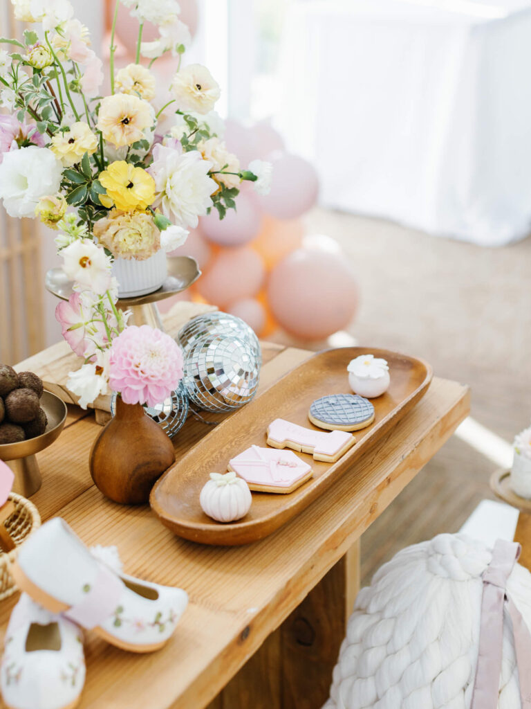 Dohl party table featuring floral centerpiece with yellow and pink flowers, decorative cookies, mini pumpkins, and disco balls on a wooden surface.